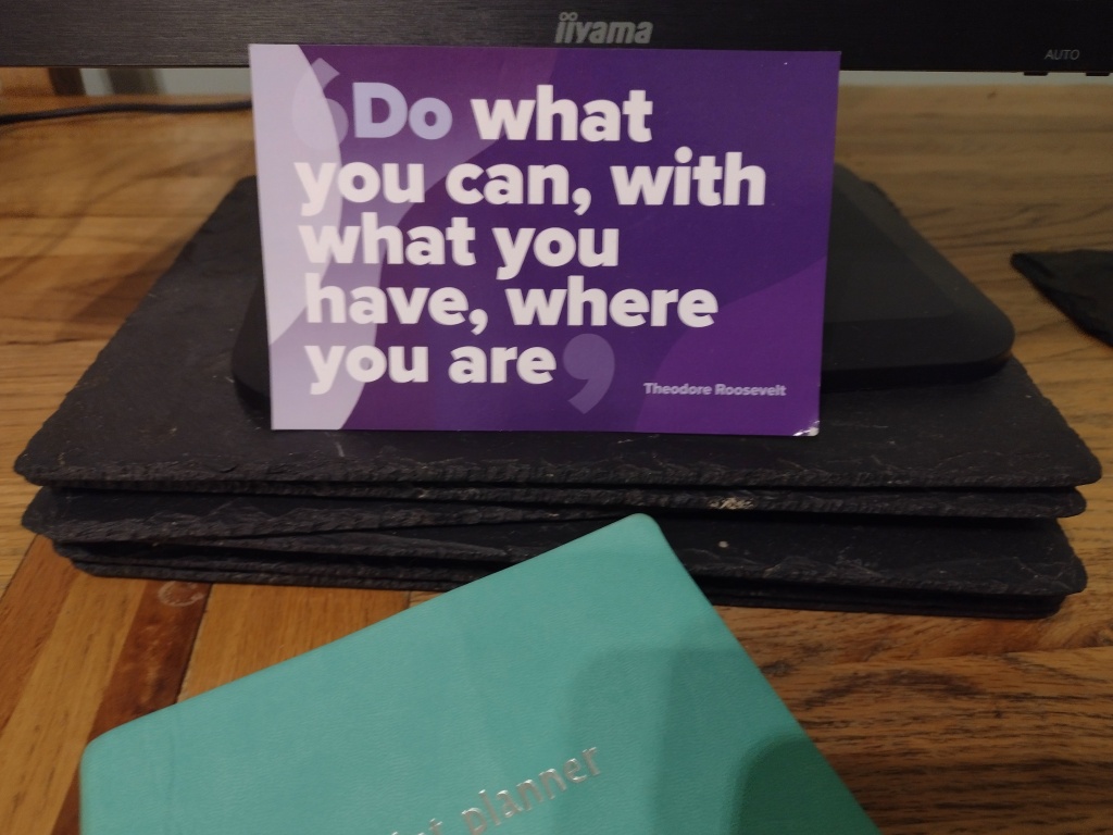 Postcard saying 'Do What you can, with what you have, where you are' by Theodore Roosevelt.  The postcard leans on a computer screen